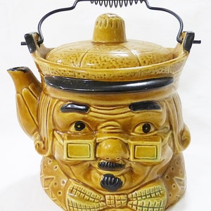 Puede incluir: Una tetera de cerámica marrón con forma de cara de hombre con bigote negro y gafas. La tetera tiene un asa negra y una tapa con un patrón marrón y negro.