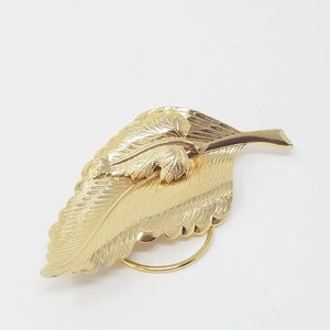 Può includere: Un fermaglio a forma di foglia dorata con un design dettagliato. Il fermaglio è in metallo e ha uno stile semplice ed elegante.
