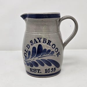 May include: Gray ceramic pitcher with a dark blue rim, handle, and decorative leaf design. The text "OLD SAYBROOK EST. 1635" is printed on the front. The pitcher has a spout for pouring.