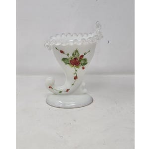 Puede incluir: Jarrón de vidrio blanco con forma de cornucopia, con un borde con volantes y una forma curva, similar a un cuerno. El jarrón está decorado con rosas rojas y hojas verdes pintadas a mano. La base es redonda y plana.