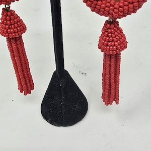 May include: Red beaded earrings with a teardrop shape and a tassel. The earrings are made of small, round beads and are displayed on a black stand against a white background. The earrings are a vibrant red colour.