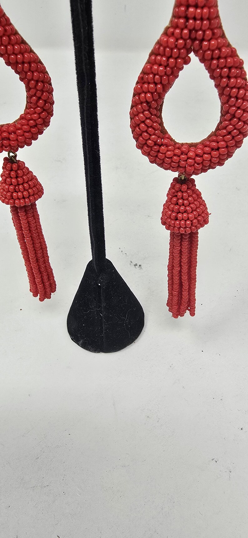 May include: Red beaded earrings with a teardrop shape and a tassel. The earrings are made of small, round beads and are displayed on a black stand against a white background. The earrings are a vibrant red colour.