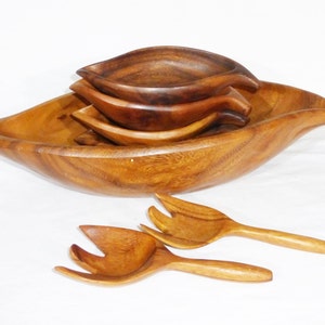 Pode incluir: Um conjunto de quatro tigelas de madeira em forma de folha, com dois talheres de salada de madeira correspondentes. As tigelas são aninhadas umas nas outras, com a tigela maior na parte inferior e a menor na parte superior. As tigelas são feitas de madeira marrom claro e têm um acabamento liso e polido.