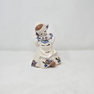 Puede incluir: Figura de cerámica de una niña holandesa con vestimenta tradicional. La figura es principalmente blanca con detalles florales azules y morados en el vestido y el sombrero. La niña está sosteniendo sus manos.