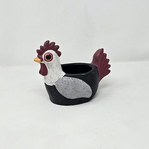 May include: A ceramic chicken-shaped planter. The planter is black with a white and gray breast, a red comb and tail, and a yellow beak. The planter is designed to hold small plants or other decorative items.