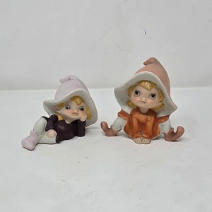 Pode incluir: Duas figuras de cerâmica de elfos. Um elfo está deitado, usando uma camisa roxa, calças brancas e um chapéu lilás claro. O outro elfo está sentado, usando um vestido laranja e um chapéu rosa. Ambos têm cabelo loiro e estão sorrindo.