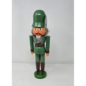May include: A wooden nutcracker soldier in a green uniform and hat. The nutcracker has a gray beard and mustache, with blue eyes and orange cheeks. The base and boots are brown.