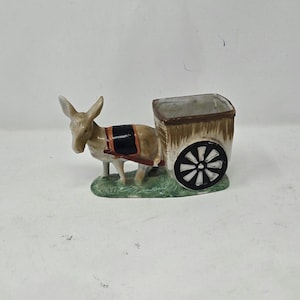 Puede incluir: Figura de cerámica de un burro tirando de un pequeño carro. El burro es de color canela con un arnés negro y marrón. El carro es marrón con una rueda de radios en blanco y negro. La figura está sobre una base verde.
