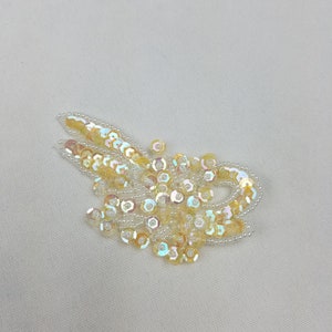 May include: A close-up of a white fabric with a design of iridescent sequins and small white beads in the shape of a swirl.