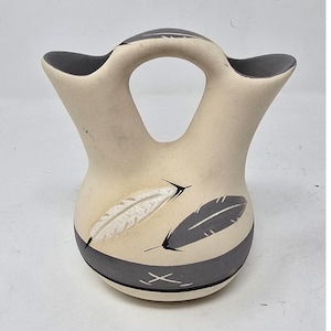 May include: A cream-colored ceramic wedding vase with two spouts connected by a central handle. The vessel features a dark gray band at the base and around the inner rims, decorated with painted white and dark gray feather motifs on the body.