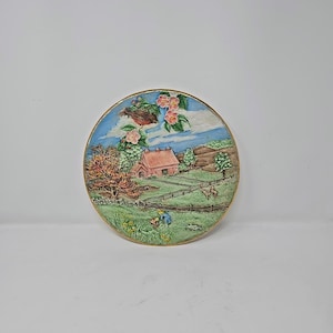 Puede incluir: Un plato decorativo redondo con una escena de paisaje rural. El plato presenta una casa, árboles, una valla y figuras en un campo. El plato tiene un borde dorado y está pintado con varios colores.