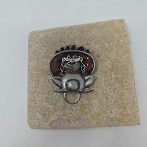 May include: A silver-colored frog-shaped pin with a hat and glasses. The frog has a red gem necklace and a gold-colored ring attached to its belly. The pin is on a light brown, textured surface.