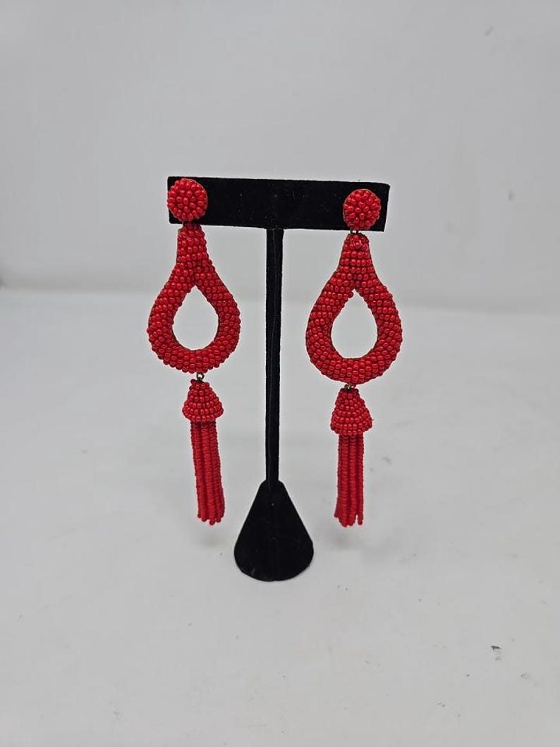 May include: A pair of red beaded earrings with a teardrop design and tassel detail. The earrings are displayed on a black stand against a white background. The earrings have a post backing.