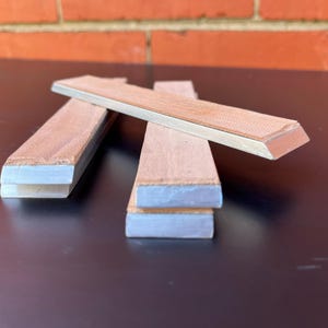 Puede incluir: Tres piedras de afilar rectangulares con una superficie marrón claro y texturizada y una base gris claro están dispuestas sobre una superficie oscura. Una piedra está en ángulo, las otras están apiladas. El fondo presenta una pared de ladrillos.