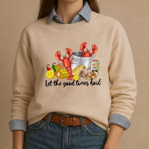 May include: Beige sweatshirt with a graphic of a crawfish boil. The design includes red crawfish, a pot, corn, potatoes, garlic, lemons, and the text "Let the good times boil". The sweatshirt has a crew neck and long sleeves.