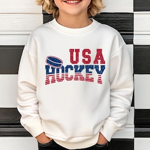 May include: A white sweatshirt with the text "USA HOCKEY" in red and blue, with a hockey puck graphic. The sweatshirt is a crew neck style.