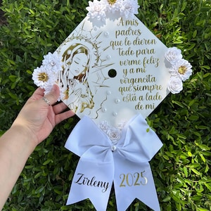 May include: White graduation cap decorated with white flowers, gold glitter, and a white bow. The cap features a gold image of the Virgin Mary and the text "A mis padres que lo dieron todo para verme feliz y a mi virgencita que siempre está a lado de mi". The bow has the text "Zirleny 2025" in gold.