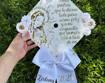 May include: White graduation cap decorated with white flowers, gold glitter, and a white bow. The cap features a gold image of the Virgin Mary and the text "A mis padres que lo dieron todo para verme feliz y a mi virgencita que siempre está a lado de mi". The bow has the text "Zirleny 2025" in gold.