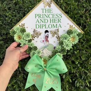 May include: A green and gold graduation cap decorated with flowers, butterflies, and a princess figure. The cap has the text "The Princess and Her Diploma" and a green bow with the text "Class of 2025".
