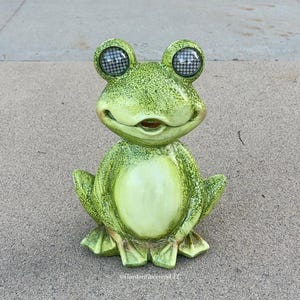 May include: A green frog figurine with a textured, mottled finish and a smooth, lighter green belly. The frog has large, prominent eyes featuring a dark, grid-like pattern. The text GardenGreeters LLC is printed in small white letters at the base.