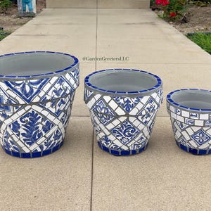 Puede incluir: Tres macetas de cerámica con diseño de mosaico. Las macetas son blancas con patrones florales y geométricos azules. Los bordes y las bases son azules. Las macetas son de diferentes tamaños.