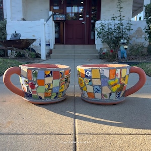 May include: Two large, mosaic-tiled teacup planters with terracotta-colored handles and rims. The mosaic tiles are in various colors and patterns, including flowers and butterflies. The planters are sitting on a concrete surface.