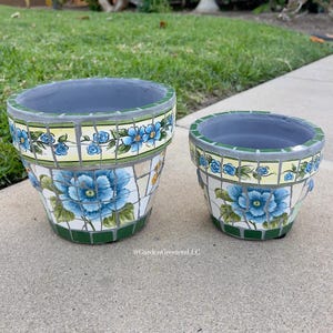 Op de afbeelding: Twee bloempotten met mozaïektegels. De grotere pot heeft een groot blauw bloemmotief, terwijl de kleinere een vergelijkbaar ontwerp heeft. Beide potten hebben een groen-gele bloemenrand en een grijze binnenkant. De potten staan op een betonnen ondergrond.