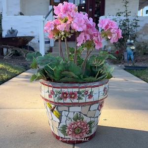 Puede incluir: Una maceta de estilo mosaico con un arreglo floral rosa. La maceta presenta un fondo blanco con detalles en rojo y diseños florales. La maceta tiene una libélula y un gran diseño de flor rosa. El texto "@GardenGreetersLLC" es visible.