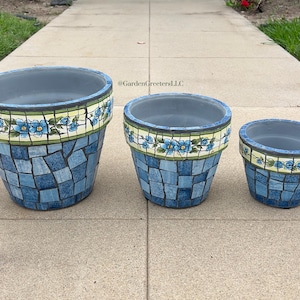 Puede incluir: Tres macetas de estilo mosaico de diferentes tamaños. Las macetas son principalmente azules con un patrón de mosaico y una banda decorativa de flores azules y hojas verdes. Las macetas están dispuestas sobre una superficie de hormigón.
