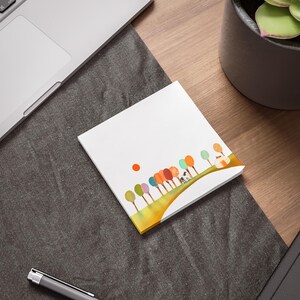 May include: A white notepad with a colorful illustration of trees, a sun, and a small house. The trees are various colors, including green, orange, and blue. The notepad is on a wooden surface next to a laptop and a pen.