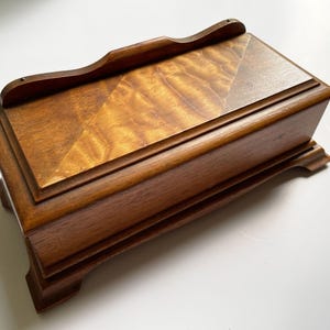 Może przedstawiać: Prostokątne drewniane pudełko z otwieraną pokrywą i dekoracyjną podstawą. Pokrywa ma wzór rombu i zakrzywiony uchwyt. Drewno ma ciepły, brązowy odcień z widocznym usłojeniem. Przedmiot do przechowywania.