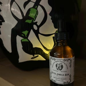 Op de afbeelding: Een bruine glazen fles met een druppelaar, met het label "Bitter Vanilla Bean" en "Aromatic Essence". Het etiket bevat een logo en tekst. Achter de fles een zwart-wit geschilderd ontwerp met een groene fles.