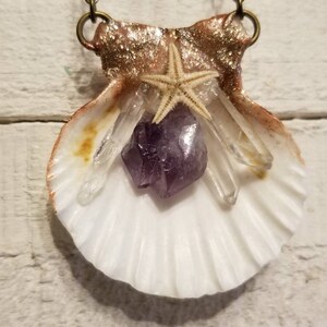 May include: A white seashell pendant necklace with a gold-toned chain. The seashell is decorated with a purple amethyst crystal, clear quartz crystals, and a white starfish.
