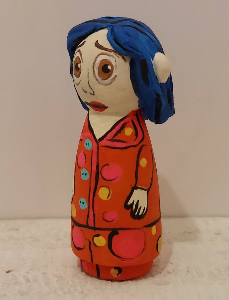 Puede incluir: Mu&ntilde;eca de madera pintada a mano con cabello azul, un vestido de lunares rojos, rosas y amarillos y una cara blanca con ojos marrones.