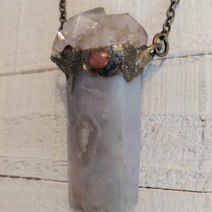 May include: A silver chain necklace with a large, clear quartz crystal pendant. The crystal is wrapped in a gold-toned metal band with a small, pink gemstone in the center.