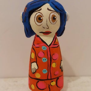 Puede incluir: Una mu&ntilde;eca de madera pintada a mano de una ni&ntilde;a con cabello azul, vestida con un vestido de lunares rojos y blancos con botones amarillos y azules.