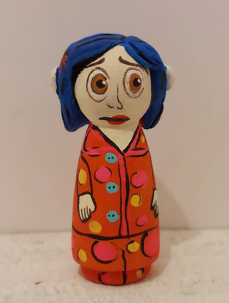 Puede incluir: Una mu&ntilde;eca de madera pintada a mano de una ni&ntilde;a con cabello azul, vestida con un vestido de lunares rojos y blancos con botones amarillos y azules.