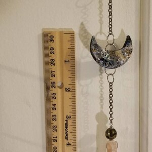 May include: A silver chain with two crescent moon charms hangs from a wooden ruler. The ruler is marked with inches and the numbers 1 through 6. The chain ends with a brown bead and a clear quartz crystal.