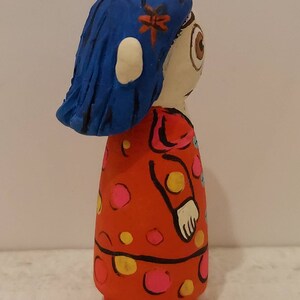 Puede incluir: Una mu&ntilde;eca pintada a mano con cabello azul, un vestido rojo con lunares amarillos y rosados, y un ojo visible.