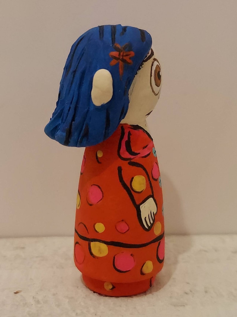 Puede incluir: Una mu&ntilde;eca pintada a mano con cabello azul, un vestido rojo con lunares amarillos y rosados, y un ojo visible.