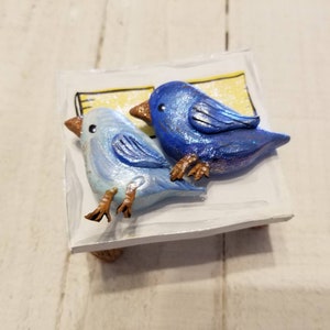 May include: Two blue birds, one light blue and one dark blue, are perched on a small white wooden shelf. The birds are made of clay and have painted details. The shelf has a yellow and white background.