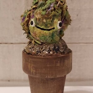 May include: A small, green, and brown potted plant with a whimsical face. The plant has large, white eyes and a small, black mouth. The pot is brown and has a rough texture.
