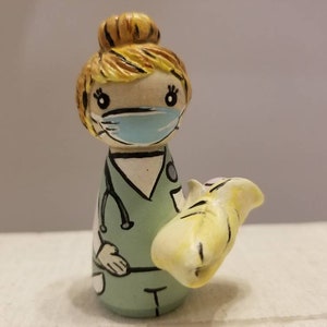 May include: A hand-painted wooden figurine of a nurse wearing a light blue medical scrubs, a blue face mask, and a stethoscope. The nurse is holding a yellow flower.