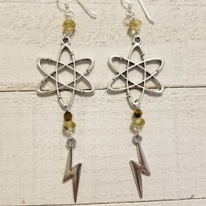 May include: Silver earrings with a lightning bolt charm and an atom symbol. The earrings have clear and amber colored crystals.