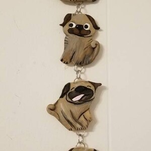 May include: A string of five pug dog ornaments hanging from a silver chain. Each ornament is made of brown clay and has a different expression. The ornaments are hanging on a white wall.