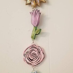 May include: A hanging floral garland with a pink rose, a yellow sunflower, a purple tulip, a pink rose, a blue hydrangea, and a bee.