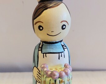 May include: A hand-painted wooden figurine of a baker wearing a white chef's hat and a blue apron. The baker is holding a green and gold cake with purple frosting and white sprinkles.