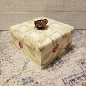 May include: A small, square, white box with a green and white floral design. The box has a brown rose on the top and pink roses on the sides. The box is sitting on a white and blue rug.