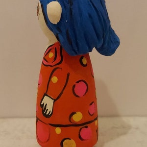 Puede incluir: Una mu&ntilde;eca de madera pintada a mano con cabello azul, un vestido rojo con lunares rosas y amarillos y manos blancas.