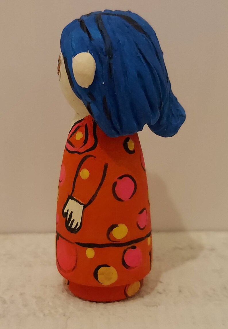 Puede incluir: Una mu&ntilde;eca de madera pintada a mano con cabello azul, un vestido rojo con lunares rosas y amarillos y manos blancas.